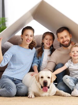 Young family with kids and dog holding roof over heads and smiling happily while sitting in living room of new apartment